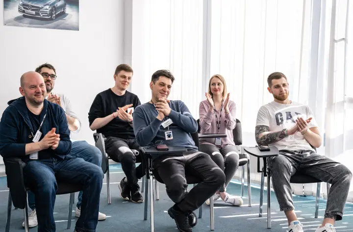 Люди сидят в ряд в комнате с плакатом на стене и дневным светом из окон.