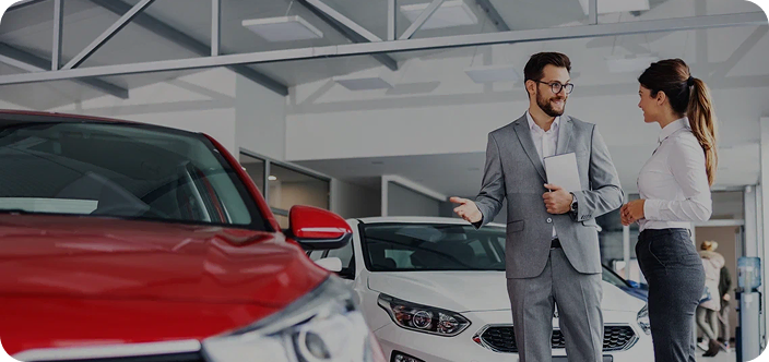 Продавец в костюме консультирует клиентку в автосалоне рядом с автомобилями.