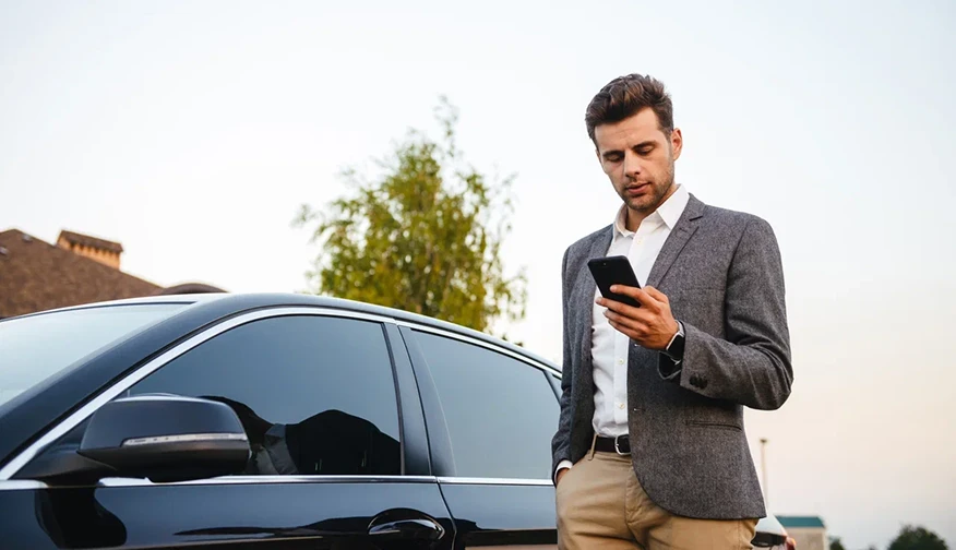 Мужчина в пиджаке стоит у чёрного автомобиля и смотрит в смартфон.