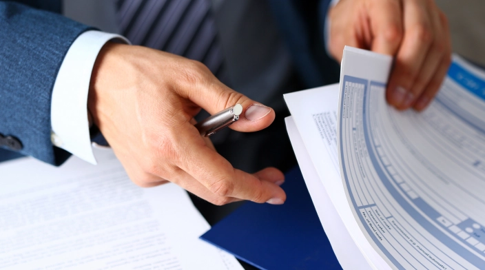 Человек в деловом костюме держит ручку и документы, внимательно изучая бумаги.