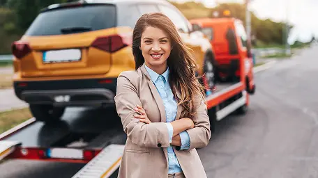 Девушка в деловом костюме стоит на фоне эвакуатора с буксируемым автомобилем.