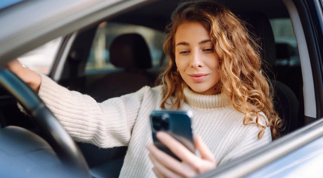 Девушка в светлом свитере управляет автомобилем, при этом её взгляд направлен на экран телефона, который она держит в руках.