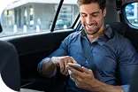 Мужчина в рубашке сидит на заднем сиденье автомобиля и улыбаясь использует смартфон.