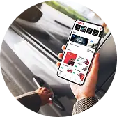 Рука держит смартфон с приложением, на экране которого отображаются машины, а на заднем плане виден реальный автомобиль